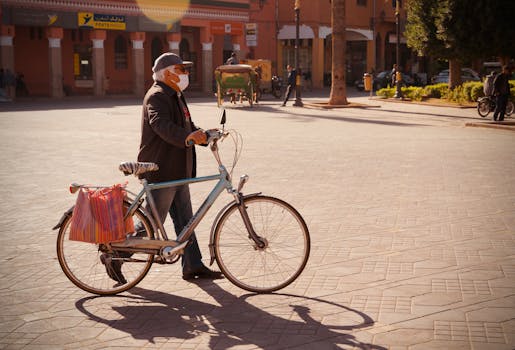Elderly man wearing a mask walking a bicycle on a sunny day in a town square.