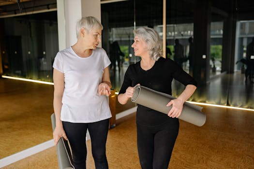 Two senior women engaging in conversation while holding yoga mats inside a fitness studio.